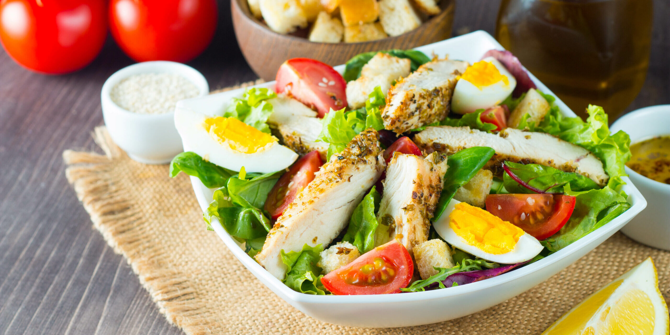Fresh Caesar salad with delicious chicken breast, ruccola, spinach, cabbage, arugula, egg, parmesan and cherry tomato on wooden background. Oil, salt and pepper. Healthy and diet food concept.