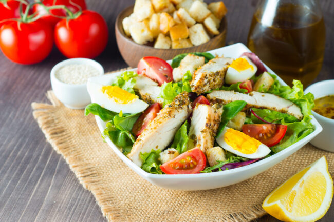 Fresh Caesar salad with delicious chicken breast, ruccola, spinach, cabbage, arugula, egg, parmesan and cherry tomato on wooden background. Oil, salt and pepper. Healthy and diet food concept.