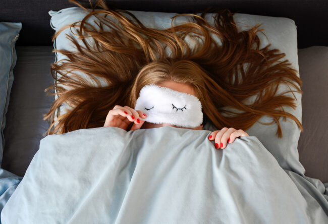 Young woman is sleeping in bed with sleep mask and peeking from