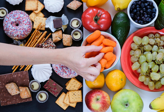 Healthy and unhealthy food concept, fruits and vegetables vs donuts, sweets and chocolate with woman’s hand. Top view