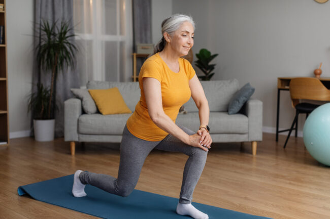 Positive sporty senior woman keeping fit at home, doing lunges, exercising on yoga mat in living room and smiling